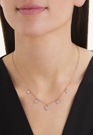 Vrouw met een gouden ketting met vijf kleine hartvormige hangers bezet met diamanten, gekleed in een zwarte top.