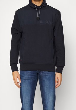 Man draagt een zwarte pullover met capuchon met de tekst "Hilfiger" en blauwe jeans, staand tegen een effen witte achtergrond.