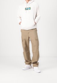 Vit hoodie med ett centrerat grafiskt tryck i blått och leopardmönstrade accenter, tillsammans med ljusbruna cargo byxor och vita sneakers.