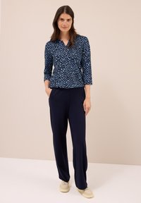 Mujer de pie con las manos en los bolsillos, vistiendo una blusa de manga larga con estampado azul, pantalones de pierna ancha azul marino y zapatillas beige sobre un fondo neutro.