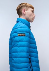 Giacca a vento blu con colletto alto, caratterizzata da una texture trapuntata orizzontale, logo a patch nero sulla manica superiore e zip laterali.