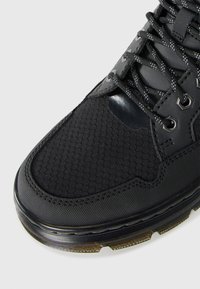 Botte noire texturée avec tissu en maille hexagonale, lacets noirs avec points réfléchissants et semelle robuste translucide sur fond clair.