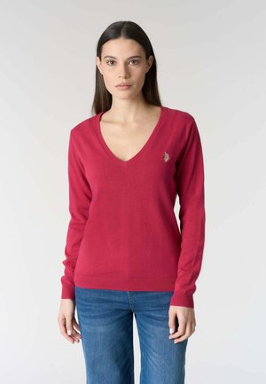 Donna con lunghi capelli castani che indossa un maglione rosso scollo a V e jeans blu, in piedi contro uno sfondo chiaro e uniforme.