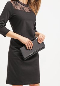 Czarna sukienka z koronkowym dekoltem, długimi rękawami i dopasowanym krojem. Trzyma czarną kopertówkę z metalicznym akcentem. Gładka faktura, elegancki design.