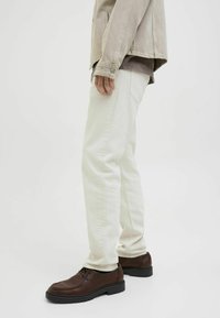 Jeans beige chiaro con una vestibilità dritta, abbinati a scarpe marrone scuro. Il tessuto sembra robusto con dettagli minimi visibili.