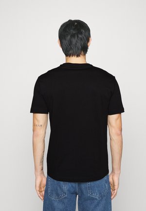 Mann mit kurzen schwarzen Haaren trägt ein schlichtes schwarzes T-Shirt und blaue Jeans, steht mit dem Rücken zur Kamera und zeigt Tätowierungen am rechten Arm.