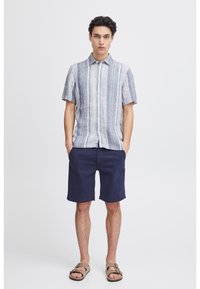 Chemise à manches courtes avec des rayures verticales bleues et blanches, associée à un short noir marine et des sandales marron. Tissu lisse, design avec col.