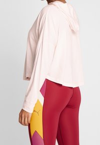 Persoon in een lichtroze, met capuchon, lange mouw top en rode leggings met gele, roze en witte geometrische panelen en het Puma-logo aan de zijkant.