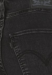 Bolsillo de unos vaqueros de mezclilla negra con diseño arcuado cosido, etiqueta de tela de Levi's visible y parche de cuero parcialmente visible con el logo del caballo.
