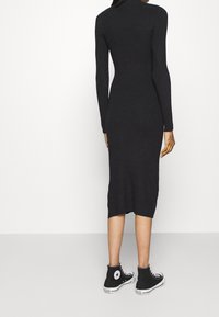 Femme portant une robe midi noire à manches longues côtelées et ajustée ainsi que des baskets montantes Converse noires, debout devant un fond blanc.