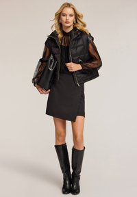 Schwarze gepolsterte Weste mit Kapuze, durchsichtige Spitzenlange Ärmel, einen schwarzen Wickelrock und kniehohe strukturierte schwarze Stiefel. Hält eine schwarze Tasche.
