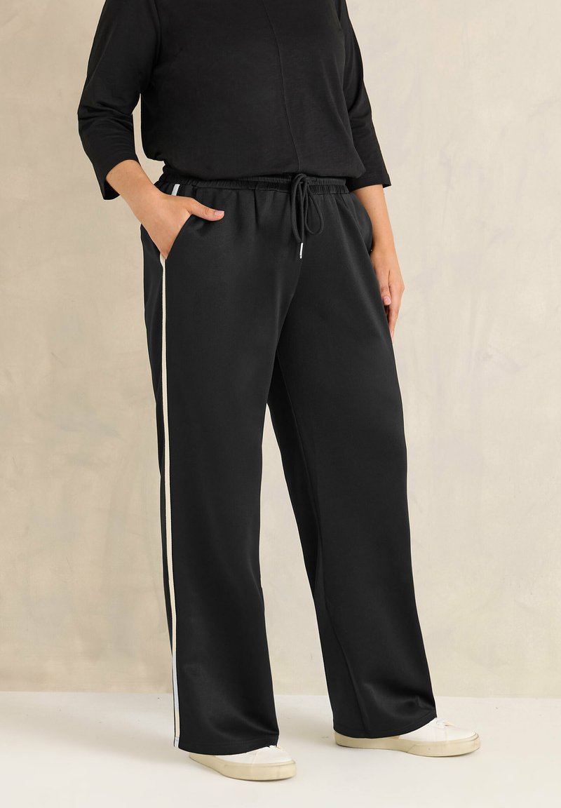 Persona vestida con pantalones anchos negros con franjas blancas laterales y una camiseta negra, de pie con una mano en el bolsillo y zapatillas blancas.