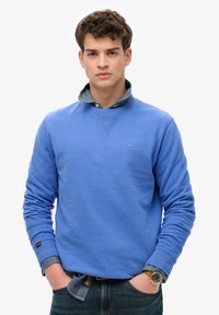 Sudadera de algodón azul con puños y cuello de canalé, superpuesta sobre una camisa de mezclilla. Presenta un pequeño logo bordado en el pecho.