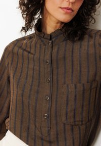 Chemise à rayures brunes avec des lignes bleues, col mandarin, fermeture à boutons à l'avant et poche poitrine. Le tissu semble texturé et subtil.