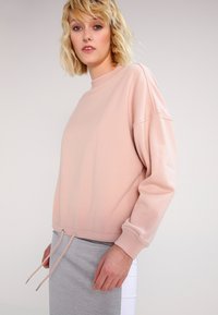 Rózsaszín pulóver hosszú ujjal, kerek nyakkivágással, állítható zsinórral az alján és bordázott mandzsettákkal. Puha anyag, laza illeszkedés.