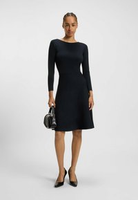 Robe noire à manches longues avec un corsage ajusté et une jupe évasée, présentant un motif texturé. Associée à des talons hauts noirs et un petit sac à main.