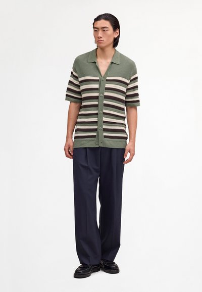Uomo che indossa una maglia a maniche corte a righe verdi e beige, pantaloni larghi scuri e scarpe nere, in piedi davanti a uno sfondo bianco uniforme.