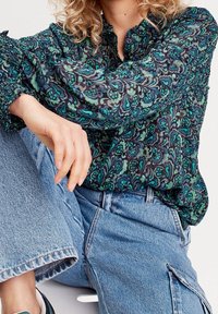 Blouse à motifs floraux en bleu foncé et vert, en tissu léger. Dotée d'un col montant et de manches élastiquées, associée à un jean en denim.