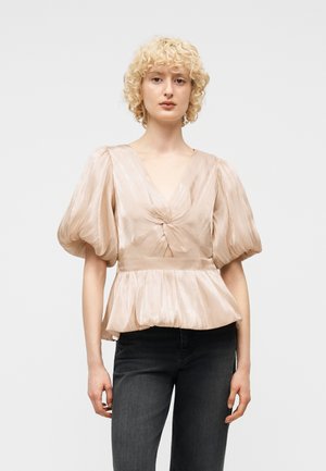 Ξανθιά γυναίκα που φοράει ανοιχτό ροζ σατέν μπλούζα με φουσκωτά μανίκια και βολάν στο τελείωμα, συνδυασμένη με μαύρο τζιν, στέκεται μπροστά σε απλό φόντο.