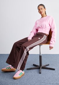 Vaaleanpunainen lyhyt collegepaita, jossa on "ADIDAS"-teksti, ruskeat leveälahkeiset housut, joissa on valkoiset raidat, sekä vaaleanpunaiset kengät vihreillä yksityiskohdilla.