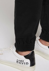 Pantaloni jogger neri con polsini arricciati, abbinati a sneakers bianche in pelle con dettagli neri e marchio stampato di lato.