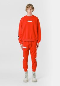 Sudadera roja y pantalones de chándal a juego presentan un parche gráfico, bolsillo con cremallera y acentos bordados en blanco, combinados con botas claras.
