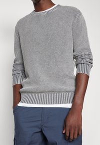 Harmaa neulepusero teksturoidulla kuviolla, jossa on raidalliset hihansuut ja helmassa. Pyöreä kaula-aukko, kerroksittain valkoisen paidan päällä.