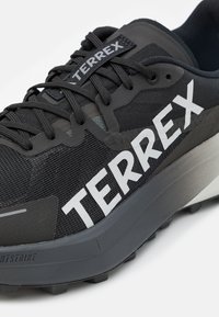 Juodas Terrex batų modelis su tinkleliu viršuje, storu pilku padu, išryškintu baltu logotipu ir tekstūruota danga, užtikrinančia sukibimą ir atramą.