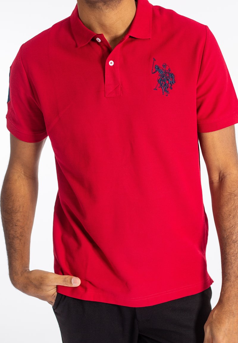 Homme portant un polo rouge vif avec un logo brodé bleu foncé de cheval et cavalier sur la poitrine, main dans la poche, sur fond blanc.