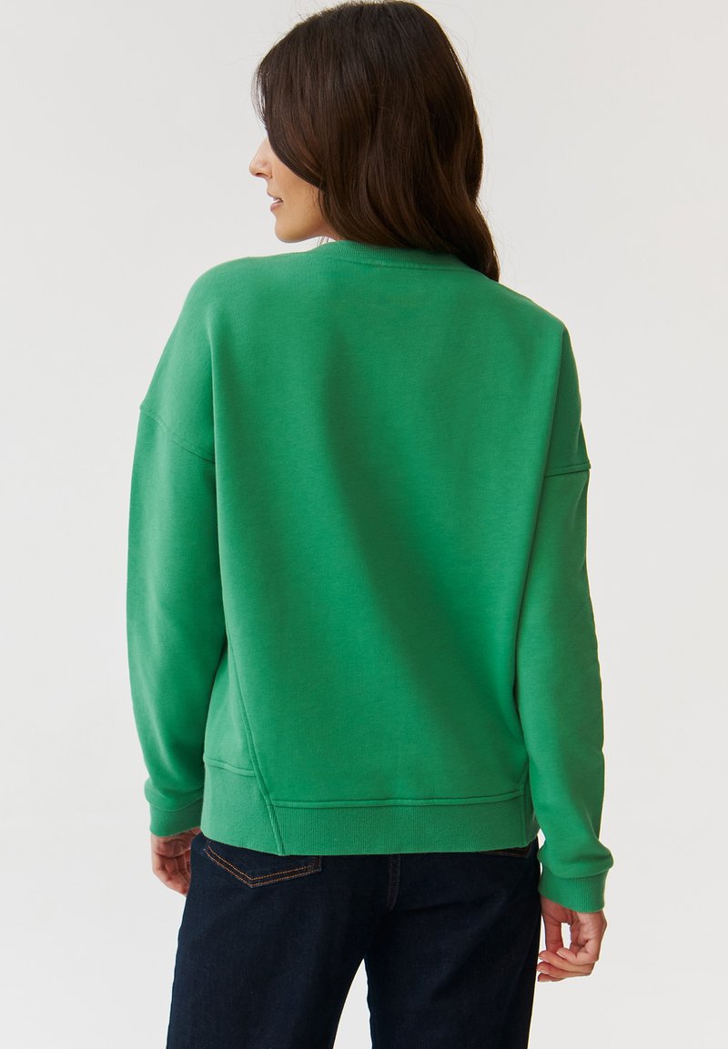 Zielona bluza z luźnym krojem, okrągłym dekoltem i prążkowanymi mankietami, w połączeniu z ciemnymi dżinsami. Tylny widok podkreśla gładką fakturę.