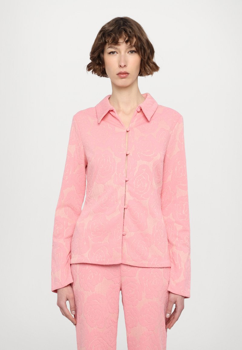 Pink floral-patterned blouse with long sleeves, button closure, and a collared neckline. Made of textured fabric.