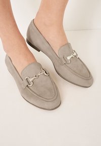 Grijze suède loafers met een spitse neus, voorzien van een zilveren metalen hardware detail over de bovenkant en een lage, platte hak.