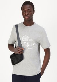 Tricou gri cu guler rotund, decorat cu un print grafic alb, purtat alături de o mică geantă neagră tip crossbody, într-un stil casual de o persoană.