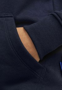 Tume sinine dressipluus, millel on tekstuuriga kude, elastsed mansetid ja esiküljel tasku, mille sees on nähtav käsi.