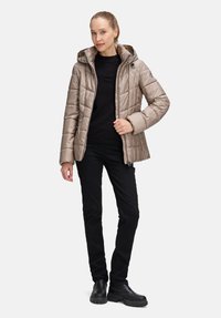 Jasnobeżowa pikowana kurtka z kapturem, zapinana na zamek z przodu, z bocznymi kieszeniami. Noszona na czarnym swetrze i czarnych spodniach.