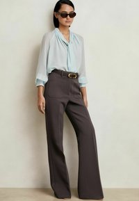 Blusa azzurra chiara con scollatura legata, abbinata a pantaloni marroni scuri a vita alta con gamba ampia e tasche, completati da occhiali da sole.