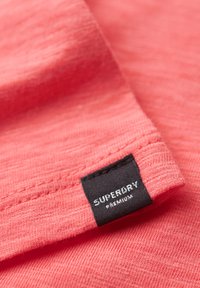 Nærbillede af koralpink stofkant med sort Superdry Premium mærkat syet på kanten.