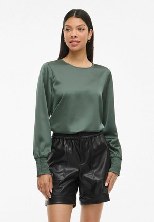 Woman wearing a satin green long-sleeve blouse with smocked cuffs and black faux leather shorts, standing against a white background.