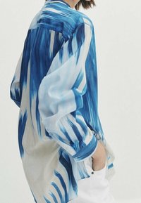 Niebiesko-biała koszula w wzory wykonana z lekkiego materiału, o luźnym kroju, z długimi rękawami i subtelnym gradientowym wzorem.