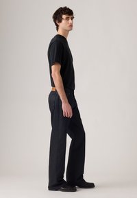 Jonge man die zijwaarts staat, gekleed in een zwart T-shirt, donkere, losse jeans en zwarte schoenen tegen een effen lichte achtergrond.