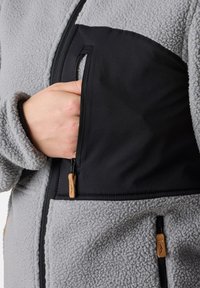 Fleece jack met grijze textuur, voorzien van een zwarte ritszak. Lederen trekband aan de rits, zachte stof met een relaxte fit.