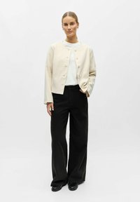 Femme debout avec une main dans la poche, portant une veste crème boutonnée, une chemise blanche, un pantalon large noir et des chaussures noires sur un fond blanc.