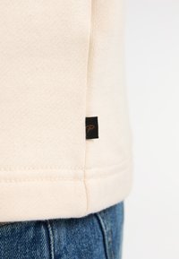 Sudadera color beige claro con bordes de canalé y un dobladillo cosido visible. Incluye una pequeña etiqueta negra con un logotipo en el lateral.