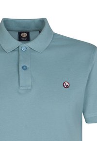 Polo de color azul claro en tela texturizada, que presenta una tapeta de tres botones y un pequeño logo circular en el lado izquierdo del pecho.