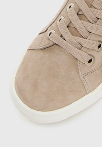 Primo piano di una punta di sneaker in suede beige con lacci coordinati e suola in gomma bianca su sfondo bianco.
