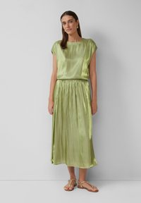 Robe en satin vert à manches courtes, taille froncée et jupe longueur cheville. Associée à des sandales plates beiges et des boucles d'oreilles en or simples.