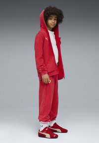Roter Sweatshirt und Sweatpants aus weichem Material, mit einem weißen Logo und einem gelben Akzent. Rote Sneakers mit cremefarbenen Details und weißen Socken.
