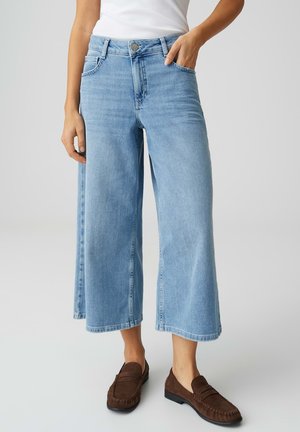 Femme portant un jean bleu clair à jambes larges et raccourci, un haut blanc et des mocassins marron, debout avec une main dans la poche, sur un fond uni.