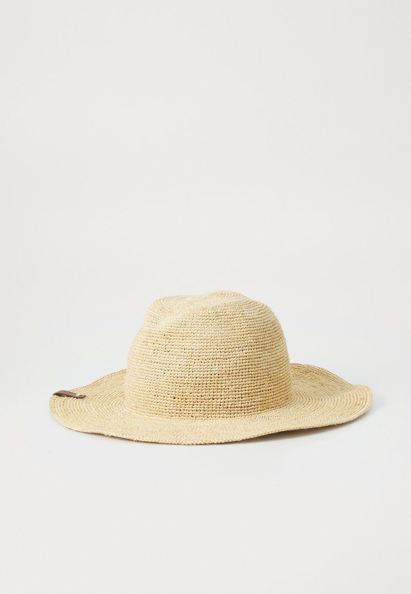 Aizaugušā salmu cepure ar plašu malumu, gaiši bežā krāsā, un brūna ādas akcenta sānos. Apaļa ķīļa ar teksturētu virsmu.