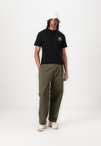 Svart bomull T-skjorte med logo, oliven grønne bukser, beige bøttehatt og lyse joggesko. Passformen er avslappet og viser frem pustende materialer.